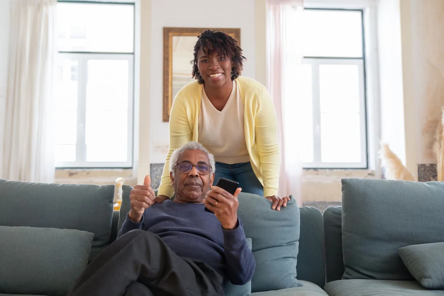 Older man holding a phone on the couch while a care worker smiles behind him