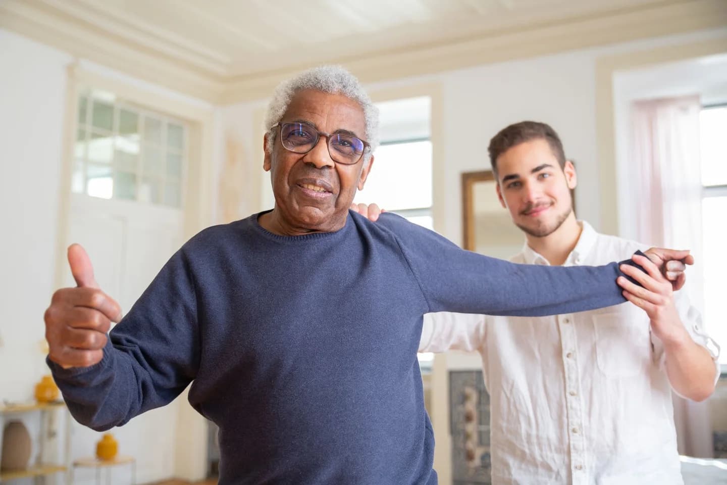 Older man smiling while a younger man supports his arm during an exercise at home
